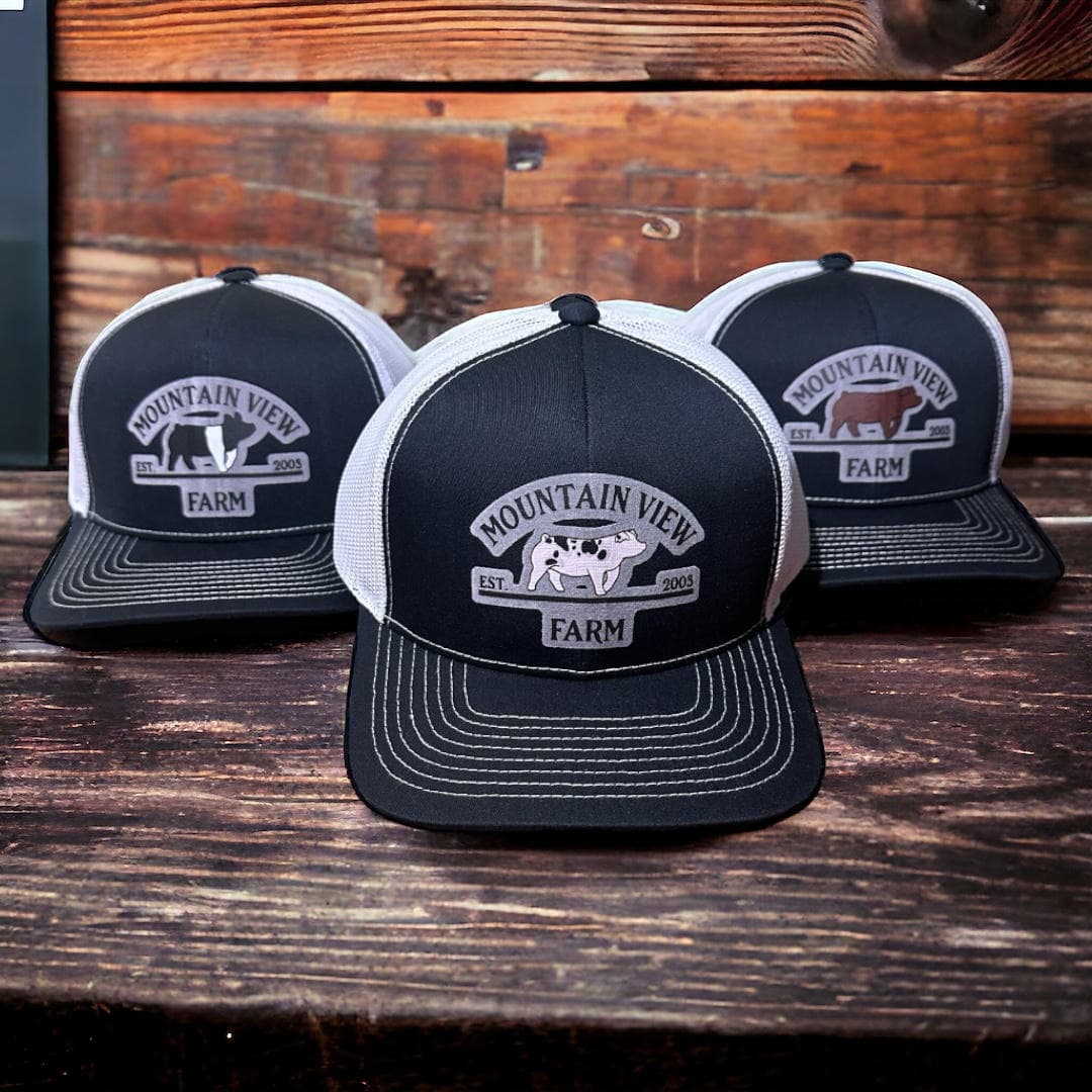Finished set of three black-and-white mesh-back trucker caps on a wood surface, each with a different Mountain View Farm animal patch centred on the black front panel.